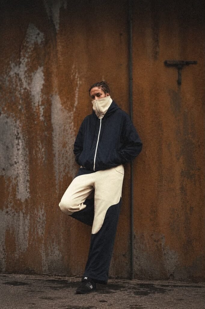 Modelo con chaqueta azul de cuello alto y pantalón con cortes curvos frente a un muro oxidado